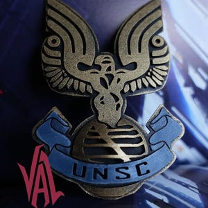 Peut inclure: Épingle à insigne UNSC de couleur bronze, représentant un aigle stylisé aux ailes déployées au-dessus d'un globe. L'emblème comporte une bannière bleue avec les lettres "UNSC", probablement un objet de collection.