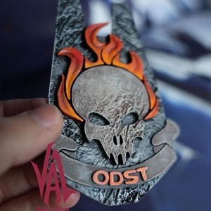 May include: A detailed, gray and orange ODST emblem featuring a skull engulfed in flames. The emblem has a textured background and the letters "ODST" are displayed on a banner. The design is held by a hand.