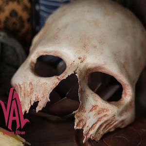 May include: A white skull mask with a broken jaw and red-brown paint accents. The mask is made of a hard material and has a realistic look.