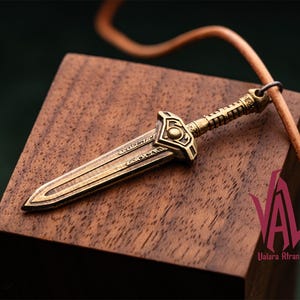 May include: A bronze-colored pendant in the shape of a sword, resting on a wooden block. The sword has intricate detailing on the handle and blade. A leather cord is attached to the pendant. The pendant is likely a piece of jewelry.