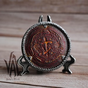 May include: A circular metal plaque with a stylised symbol and intricate details, finished in red and gold. The plaque is displayed on a silver metal stand.