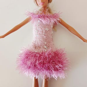 May include: A doll wearing a light pink knitted dress featuring a fuzzy off-the-shoulder neckline, a matching fuzzy hem, and scattered silver sequins across the bodice. The dress has a textured knit pattern and a vibrant pink fringe trim.