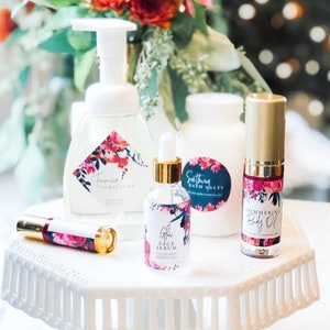 May include: A collection of beauty products, including foaming hand soap, face serum, bath salts, shimmering body oil, and a lip product, arranged on a white, tiered stand. Each product features floral designs and gold accents. The background includes a floral arrangement.