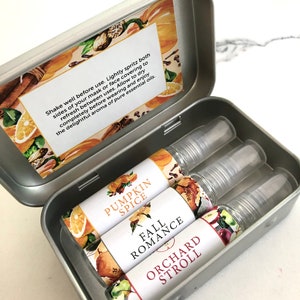 Op de afbeelding: Een zilveren metalen doos met drie essentiële olie sprays met de labels "Pumpkin Spice", "Fall Romance" en "Orchard Stroll". De binnenkant van de doos heeft een decoratieve inleg met tekst.