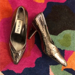 May include: A pair of silver metallic high-heeled shoes with a snakeskin pattern. The shoes have a pointed toe and a stiletto heel. The brand name "Sutton Plaza" is visible on the inside of one shoe.