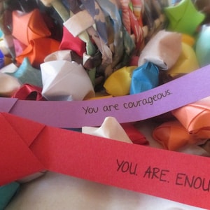 Peut inclure: Gros plan sur un tas d'étoiles en papier origami colorées avec deux bandes de papier violettes et rouges sur lesquelles sont inscrits les mots "You are courageous." et "You are enough."