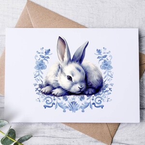 May include: A watercolor illustration of a white rabbit with blue shading, resting on a white card. The card is decorated with blue floral and scrollwork designs. The card is placed on a light wood surface with a brown envelope and green foliage.
