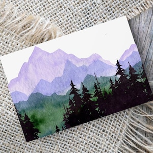 May include: Watercolor painting of a mountain landscape. The artwork features layers of purple mountains, a dark green forest, and a white sky. The painting is on a white card, resting on a burlap surface and a wooden background.
