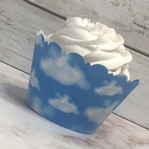 May include: A cupcake in a blue and white cloud-patterned liner. The liner has a scalloped edge and contains a swirl of white frosting. The cupcake is on a light-colored wooden surface.