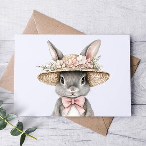 May include: A watercolor illustration of a gray bunny wearing a straw hat adorned with pink and cream-colored flowers and a pink bow tie. The bunny is centered on a white card, with a brown envelope behind it.
