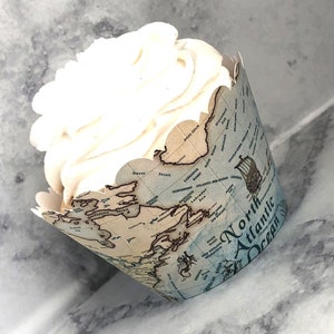 May include: A cupcake with white frosting sits in a decorative paper liner. The liner features a vintage map design with the words "North Atlantic Ocean" and illustrations of ships. The cupcake is on a marble surface.