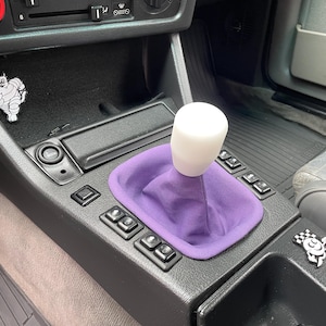 May include: A car gear shift assembly featuring a solid white cylindrical shift knob and a vibrant purple fabric shift boot. The black center console includes several rectangular buttons, a cigarette lighter socket, and two small decorative stickers.