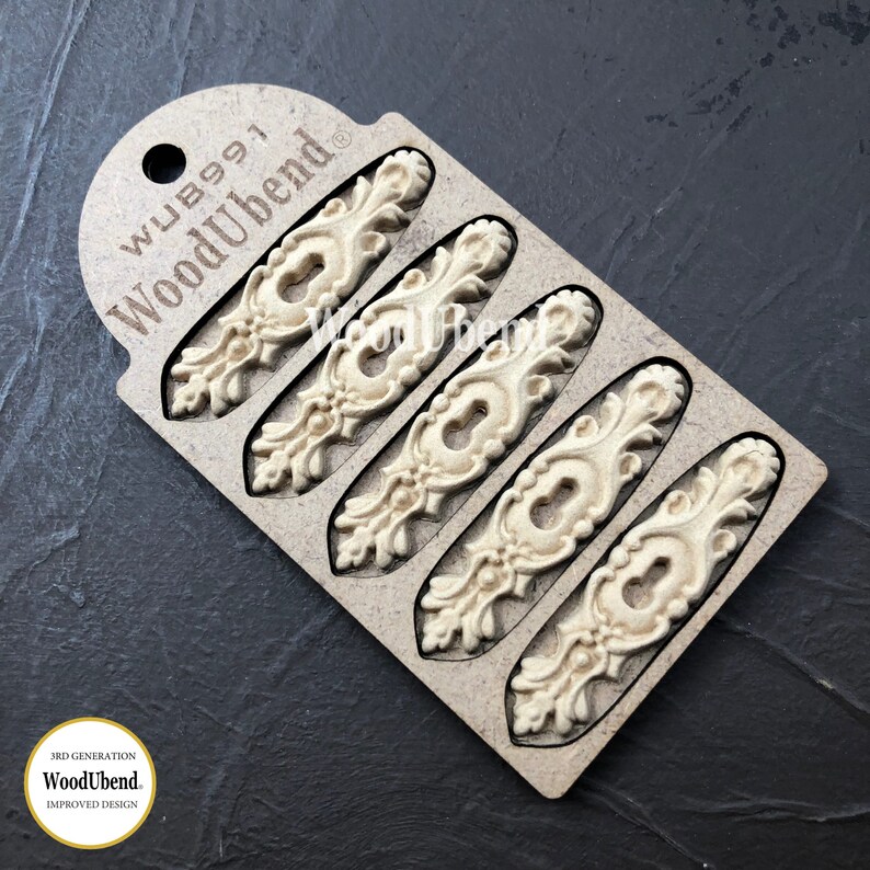 May include: A pack of five ornate, off-white wooden embellishments with a keyhole design. The embellishments are arranged in a row on a brown card with the text "WoodUbend" and "WUB991".