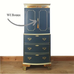 May include: A tall, dark blue cabinet with gold accents and three drawers. The cabinet has two doors with a gold tassel on the right door handle. The cabinet is on a wooden floor. WUBo991