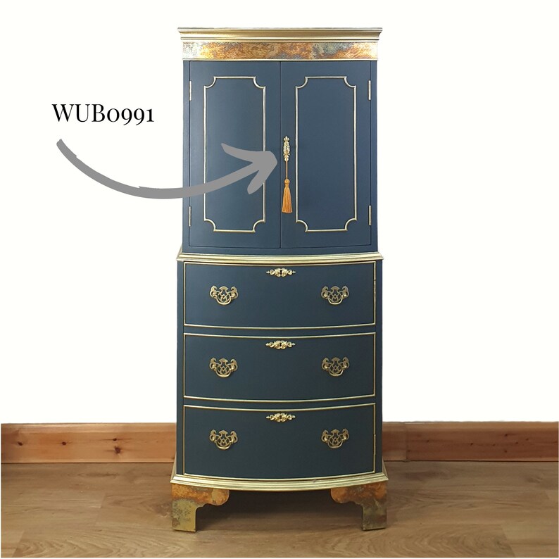 May include: A tall, dark blue cabinet with gold accents and three drawers. The cabinet has two doors with a gold tassel on the right door handle. The cabinet is on a wooden floor. WUBo991
