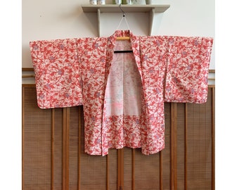 May include: A red and white patterned Japanese kimono with a floral design. The kimono is hanging on a hanger against a wooden background.