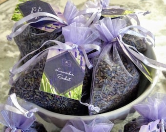 May include: A collection of lavender sachets in a white bowl. Each sachet is a small, sheer bag filled with dried lavender buds and tied with a lavender ribbon. Each sachet has a tag that reads "Lavender Sachet" with a lavender and green design.
