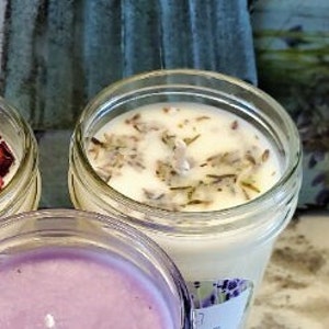 Pode incluir: Uma vela branca em um frasco de vidro transparente com flores de lavanda secas no topo. A vela tem um r&oacute;tulo com um desenho de flor de lavanda.