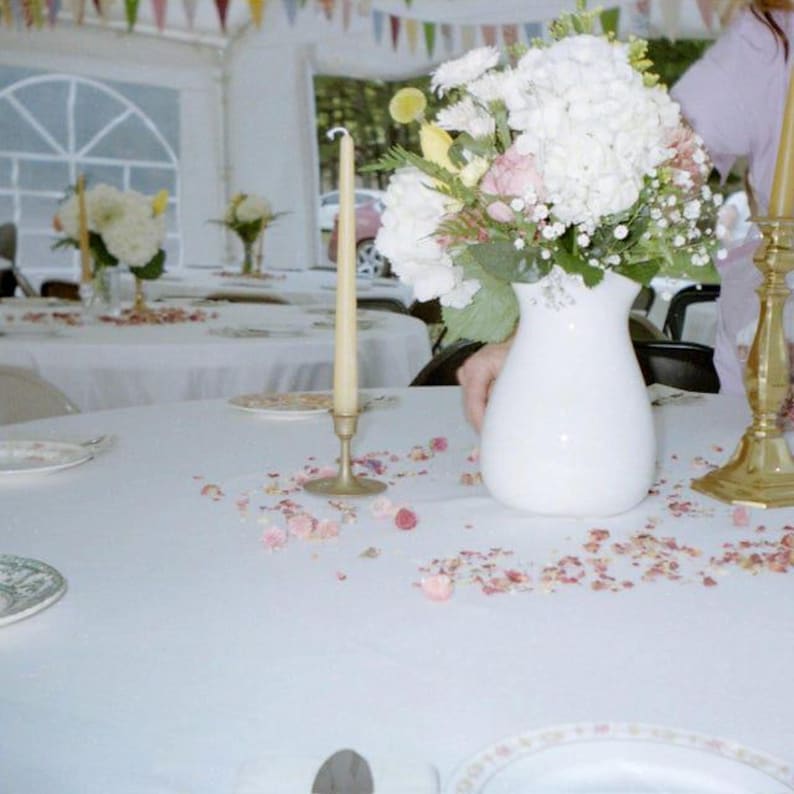 May include: A white ceramic vase filled with a bouquet of white hydrangeas, pink roses, and yellow flowers. A tall, thin, cream-colored taper candle stands in a brass holder next to the vase. Scattered pink flower petals decorate the white tablecloth.