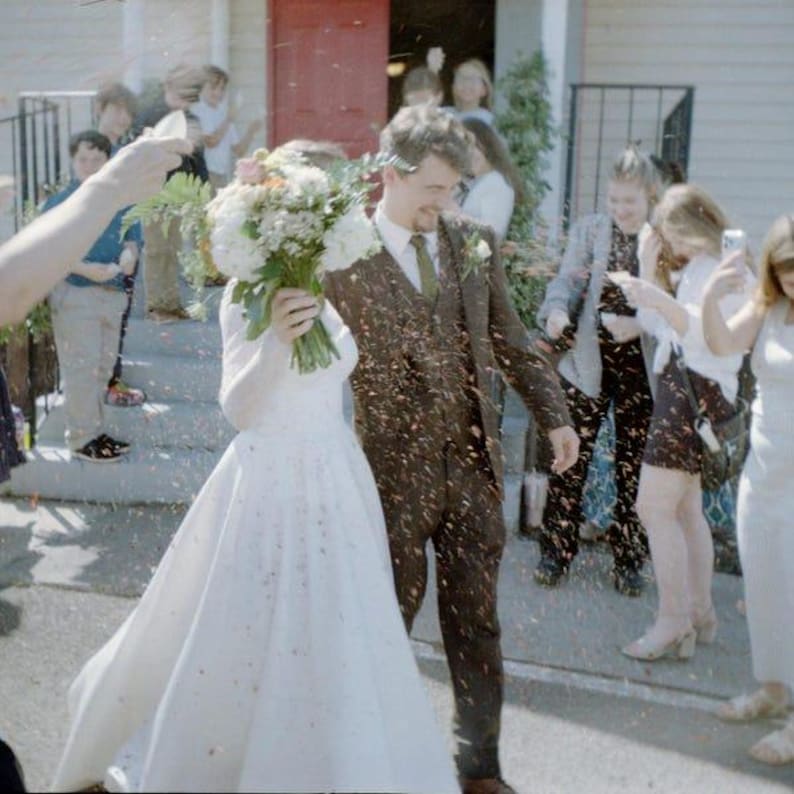 May include: A person in a white floor-length wedding dress holds a large bouquet of white and green flowers while walking next to a person in a brown three-piece suit. Confetti falls around them as they exit a building with a red door.
