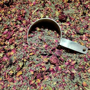 May include: A stainless steel measuring cup filled with dried lavender buds, resting on a bed of mixed dried lavender buds and vibrant pink and deep red rose petals. The metal handle of the measuring cup is visible, extending to the right side.