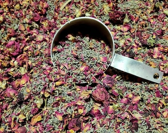 May include: A stainless steel measuring cup filled with dried lavender buds, resting on a bed of mixed dried lavender buds and vibrant pink and deep red rose petals. The metal handle of the measuring cup is visible, extending to the right side.
