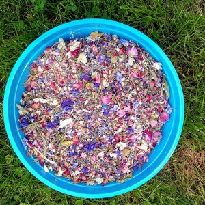 May include: A bright blue circular bowl filled with a colorful mixture of dried flower petals, including shades of pink, purple, blue, and white. The botanical blend consists of various small blossoms, buds, and stems arranged in a dense, textured pile.