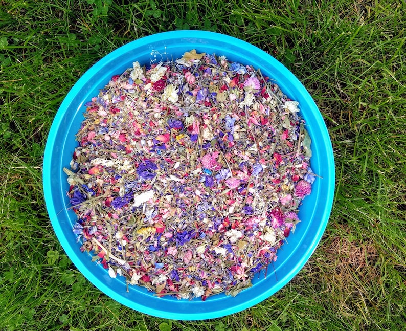 May include: A bright blue circular bowl filled with a colorful mixture of dried flower petals, including shades of pink, purple, blue, and white. The botanical blend consists of various small blossoms, buds, and stems arranged in a dense, textured pile.