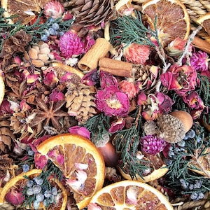 May include: A collection of dried potpourri featuring orange slices, pinecones, cinnamon sticks, star anise, pink globe amaranth flowers, dried rose petals, and green juniper sprigs with small blue berries, all arranged within a woven wicker basket.