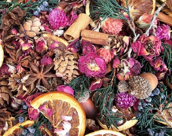 May include: A collection of dried potpourri featuring orange slices, pinecones, cinnamon sticks, star anise, pink globe amaranth flowers, dried rose petals, and green juniper sprigs with small blue berries, all arranged within a woven wicker basket.