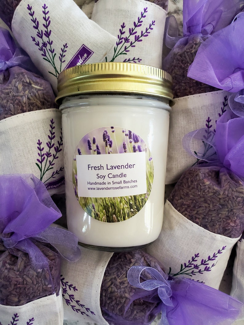 Pode incluir: Um frasco de vidro transparente com tampa dourada cheio de uma vela de soja branca. A vela tem um r&oacute;tulo redondo que diz "Fresh Lavender Soy Candle" e "Handmade in Small Batches" com o site www.lavenderrosefarms.com. A vela &eacute; cercada por pequenas bolsas de tecido roxo cheias de lavanda seca.