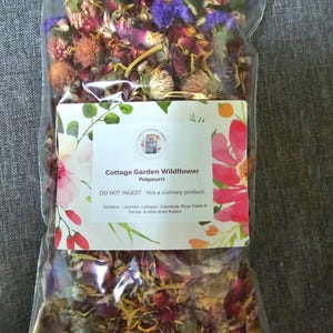 May include: A clear plastic bag filled with dried flowers, including pink, purple, and yellow petals. A white label features the text: Cottage Garden Wildflower Potpourri, DO NOT INGEST - Not a culinary product. Contains: Lavender, Larkspur, Calendula, Rose Petals & Yarrow, & other dried flowers.