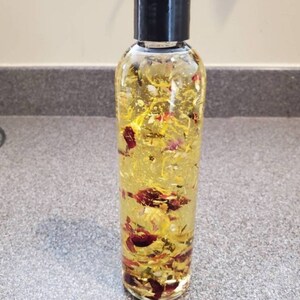 May include: A clear cylindrical plastic bottle with a black screw-top cap, filled with a translucent yellow liquid containing suspended dried flower petals in shades of red, pink, orange, and yellow, visible throughout the entire container.