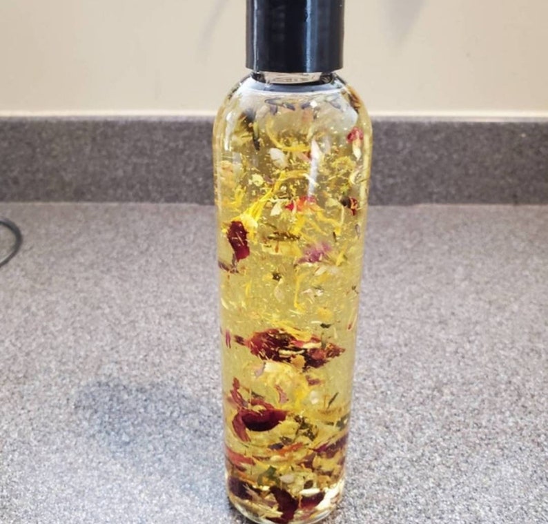 May include: A clear cylindrical plastic bottle with a black screw-top cap, filled with a translucent yellow liquid containing suspended dried flower petals in shades of red, pink, orange, and yellow, visible throughout the entire container.