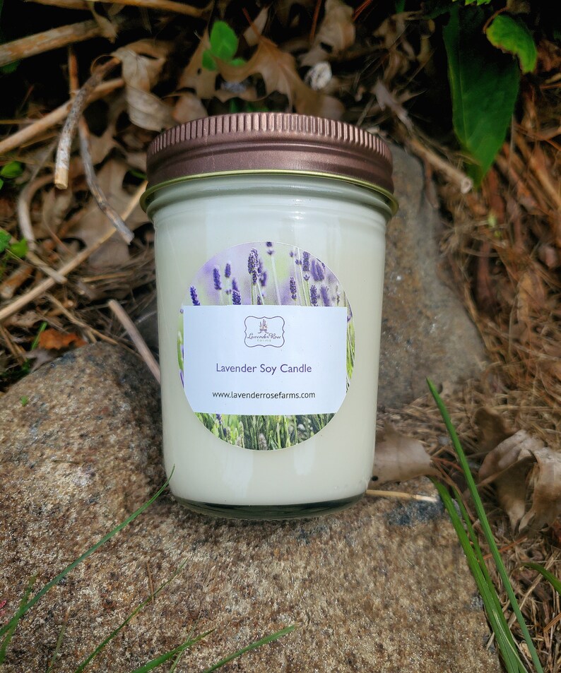 Pode incluir: Uma vela de soja branca em um pote de vidro com tampa de cobre. O r&oacute;tulo apresenta um design de lavanda e o texto "Lavender Soy Candle" e "www.lavenderrosefarms.com".