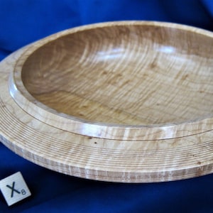 May include: A large, wooden bowl with a light brown finish. The bowl has a wide, flat base and a deep, rounded interior. The wood grain is visible throughout the bowl, and the surface is smooth and polished.