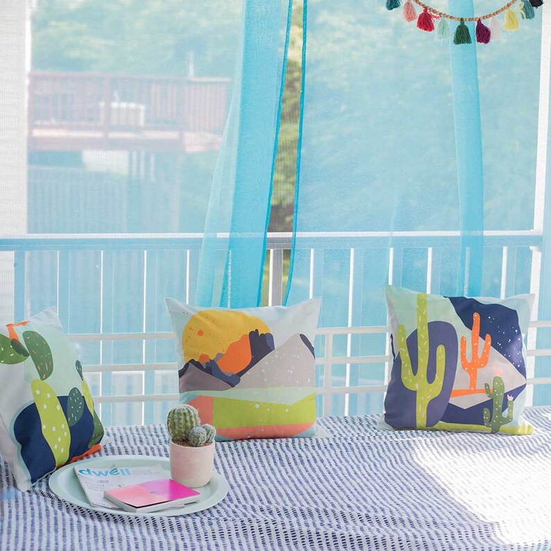 May include: Three decorative throw pillows with different patterns. The pillows are on a white and blue striped couch with a blue sheer curtain in the background. The pillows feature a desert landscape, a cactus, and a starry sky.