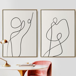 May include: Two minimalist abstract line art prints in black and white, framed in gold, hanging on a white wall. The prints feature simple, flowing lines that create a sense of movement and energy.