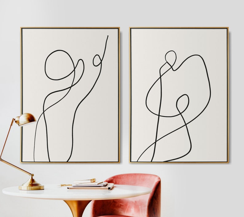 May include: Two minimalist abstract line art prints in black and white, framed in gold, hanging on a white wall. The prints feature simple, flowing lines that create a sense of movement and energy.
