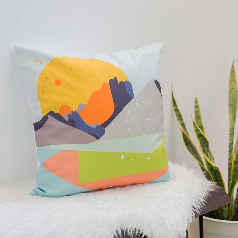 May include: A decorative pillow with a geometric design featuring a yellow sun, orange and blue mountains, and a green and orange shape on a light blue background.