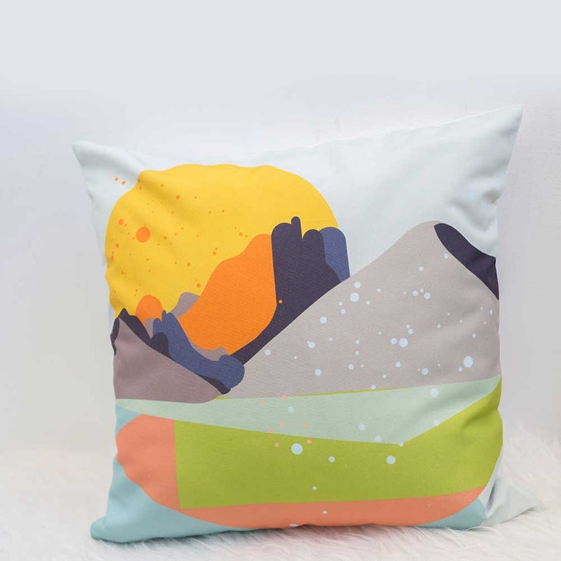 May include: A decorative pillow with a colorful abstract design featuring a yellow sun, orange and purple mountains, and a green and blue landscape. The pillow has a light blue background with white dots.