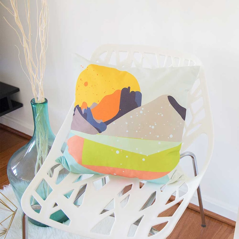 May include: A white chair with a geometric design, featuring a colorful pillow with an abstract mountain landscape and a yellow sun.