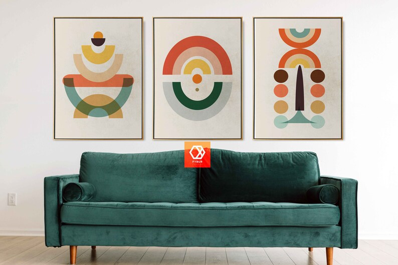May include: Three framed abstract art prints with geometric shapes in orange, yellow, teal, and brown tones. The artwork hangs above a teal velvet sofa with wooden legs. The room has a minimalist design.