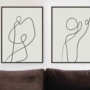 May include: Two black framed prints with abstract line art in black on a white background. The prints are hanging on a white wall above a brown couch.