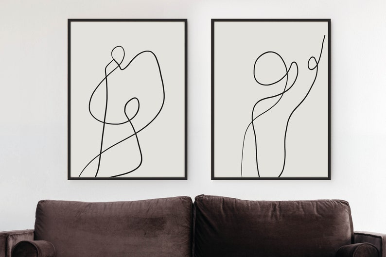 May include: Two black framed prints with abstract line art in black on a white background. The prints are hanging on a white wall above a brown couch.