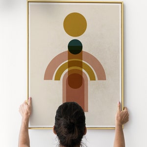 May include: A person hangs a framed abstract art print on a white wall. The print features a geometric design with circles and arches in shades of pink, orange, yellow, and green on a beige background.