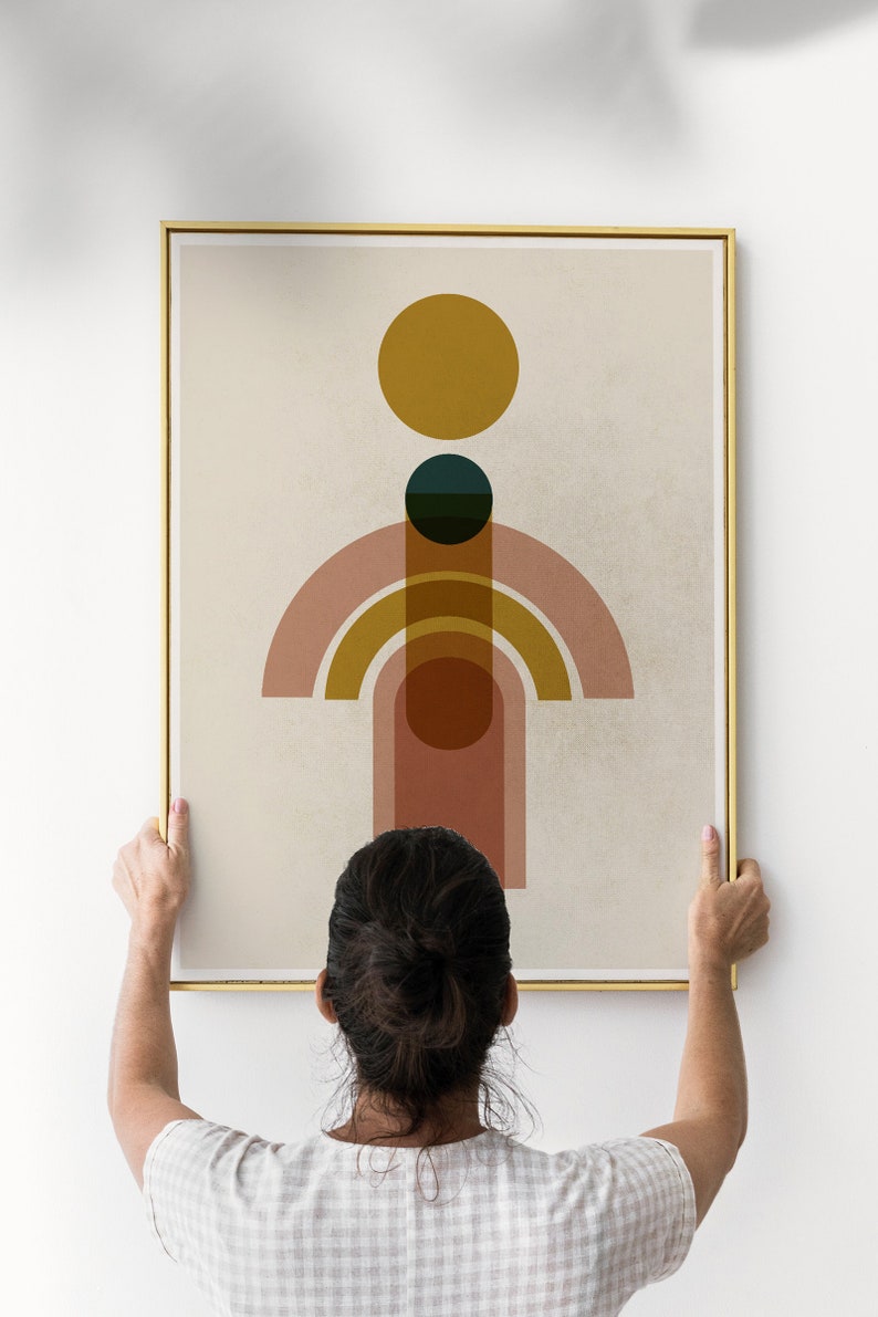May include: A person hangs a framed abstract art print on a white wall. The print features a geometric design with circles and arches in shades of pink, orange, yellow, and green on a beige background.