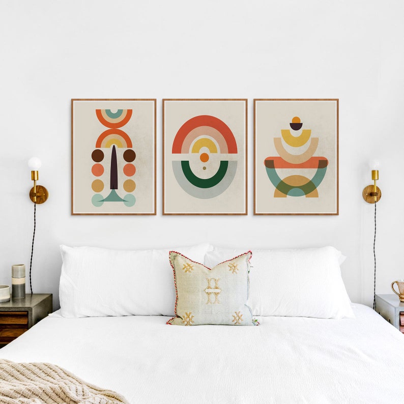 May include: Three framed abstract art prints with geometric shapes in warm colors like orange, yellow, and brown. The prints are displayed above a bed with white bedding and a decorative pillow. The frames are a light brown color.