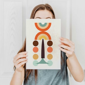 May include: Abstract art print featuring a geometric design with a cream background. The artwork includes a series of colorful shapes: orange, brown, yellow, and teal circles, and a rainbow arch. The print is held by a person.