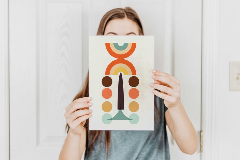 May include: Abstract art print featuring a geometric design with a cream background. The artwork includes a series of colorful shapes: orange, brown, yellow, and teal circles, and a rainbow arch. The print is held by a person.